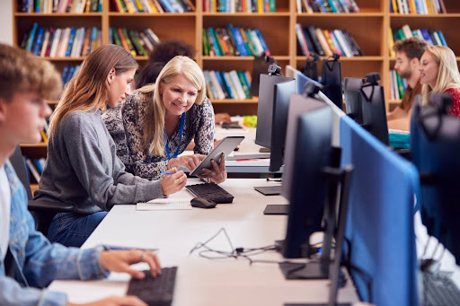 Mulher orientando aluna a realizar sua assinatura digital na biblioteca escolar.