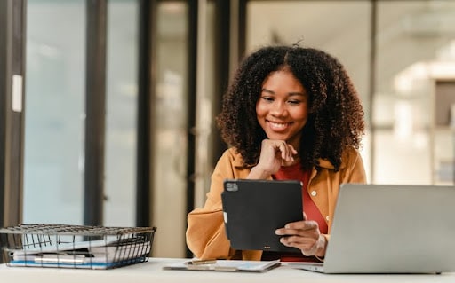 Mulher negra de frente ao notebook com tablet na mão sorrindo
